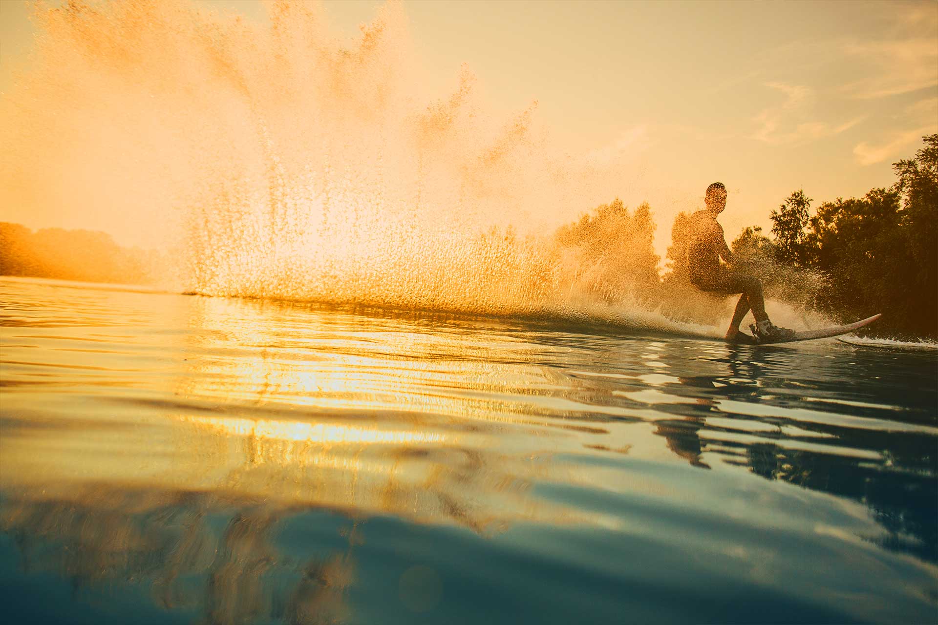 Person wakeboarding on a lake at sunset with water spraying behind them.