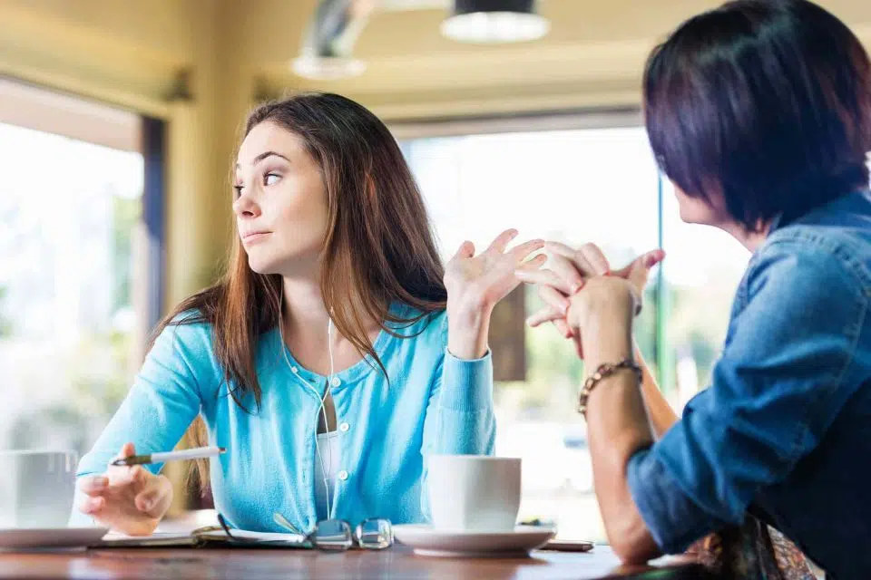 Two women sitting at a table talking while one woman ignores the other.