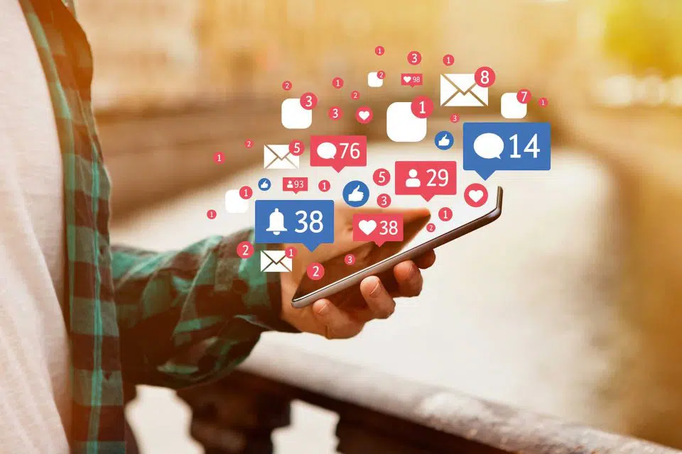 Closeup of a person's hand as they look at a smartphone with social media posting icons floating above it.